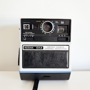 Puede incluir: Una cámara instantánea Kodak EK4 blanca y negra con una franja azul. La cámara tiene una textura de cuero negro en la parte delantera y una lente negra con un anillo plateado. La cámara tiene un dial para ajustar la distancia en metros y pies.
