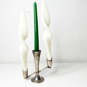 Puede incluir: Un candelero de plata con tres portavelas. Dos velas blancas están retorcidas y una vela verde está recta.