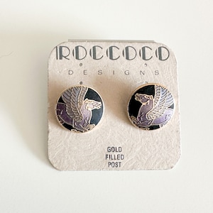 Puede incluir: Un par de pendientes de poste rellenos de oro con un diseño de esmalte negro y morado de una criatura mítica con alas. Los pendientes se muestran en una tarjeta que dice "Rococo Designs" y "Gold Filled Post".