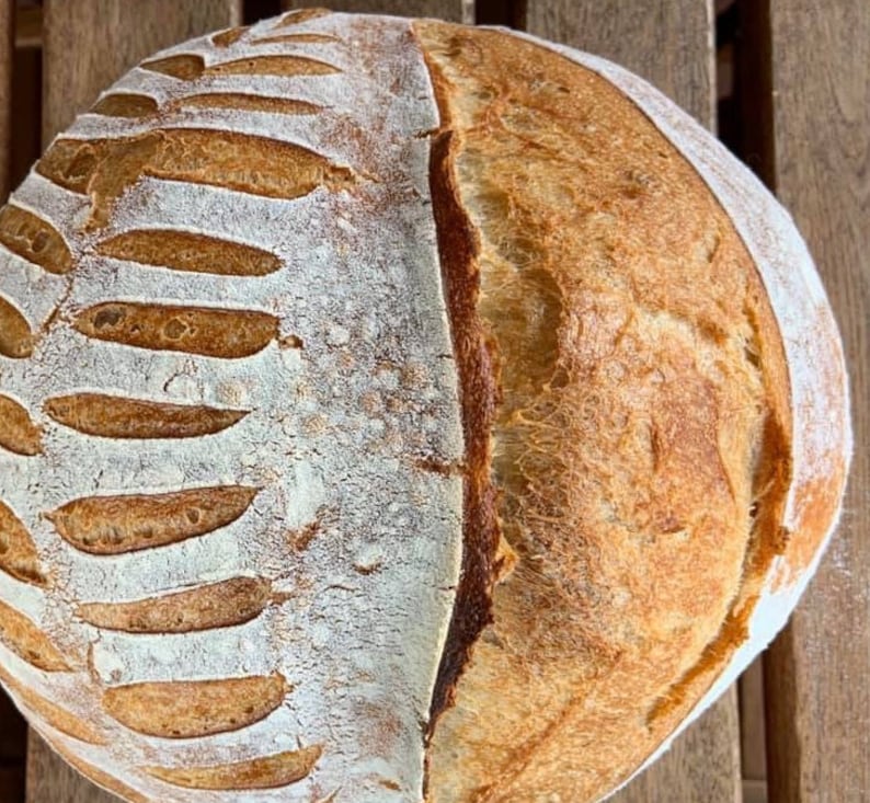 May include: A freshly baked loaf of sourdough bread with a scored crust. The bread has a light brown color and a slightly rough texture.