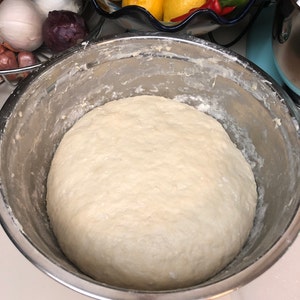 May include: A large, white dough ball sits in a stainless steel mixing bowl. The dough is smooth and slightly shiny.