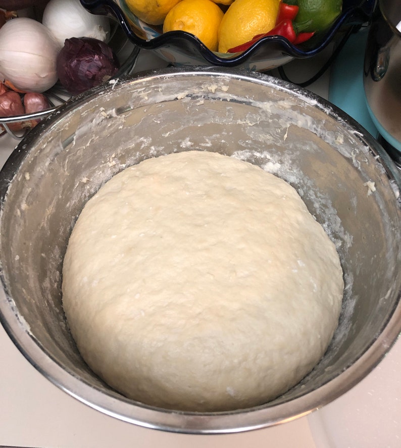 May include: A large, white dough ball sits in a stainless steel mixing bowl. The dough is smooth and slightly shiny.