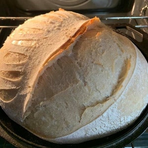 May include: A freshly baked loaf of sourdough bread with a crispy crust and a soft, airy interior. The bread is in a black cast iron pan.