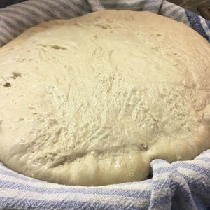 May include: A close-up of a white dough rising in a blue and white striped bowl. The dough is covered with a white cloth.