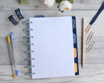 May include: A white notebook with blue and gold accents and lined pages. The notebook has a ring binder and is open to a blank page. There are two gold pens, a pencil, and paper clips on a wooden surface.