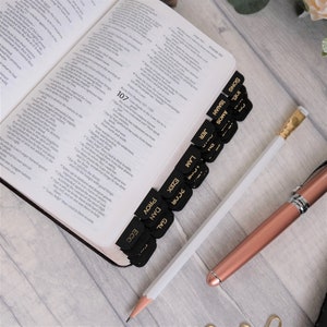 Puede incluir: Primer plano de una Biblia encuadernada en cuero negro con bordes de página dorados. La Biblia tiene pestañas negras con letras doradas para cada libro de la Biblia, incluidos los Salmos, el Cantar de los Cantares, Isaías, Amós, Lamentaciones, Ezequiel, Jeremías, Proverbios, Daniel, Eclesiastés y Gálatas. Un lápiz blanco y una pluma de color rosa dorado están sobre la Biblia.