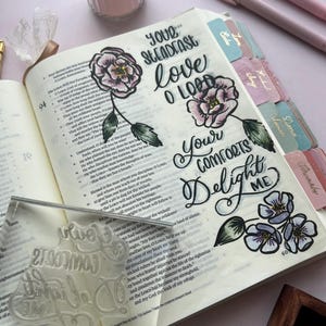 May include: An open Bible featuring floral illustrations and handwritten text, including "Your Steadfast Love O Lord" and "Your Comforts Delight Me." A clear stamp and pink candle are also visible.