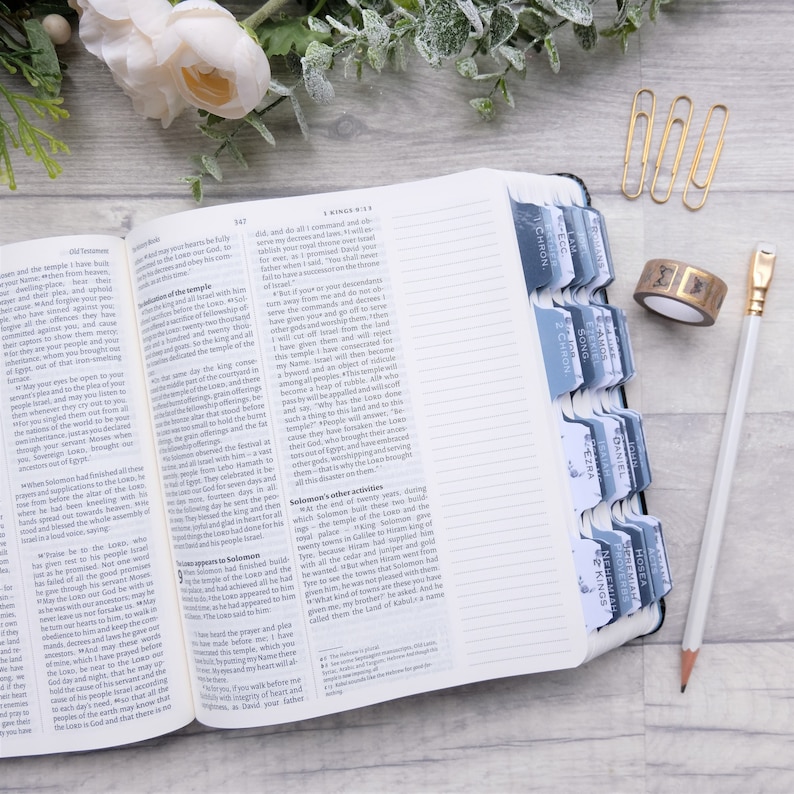 May include: A close-up of an open Bible with blue tabs marking different sections. The tabs are labeled with book names like "John", "Daniel", "Kings", and "Proverbs". The Bible is open to the book of Kings, with text visible on the pages.
