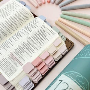 May include: Open Bible with pastel-colored page tabs and a set of 12 soft pastel highlighters. The Bible is open to a passage with text. The highlighters are arranged in a circle around the Bible.