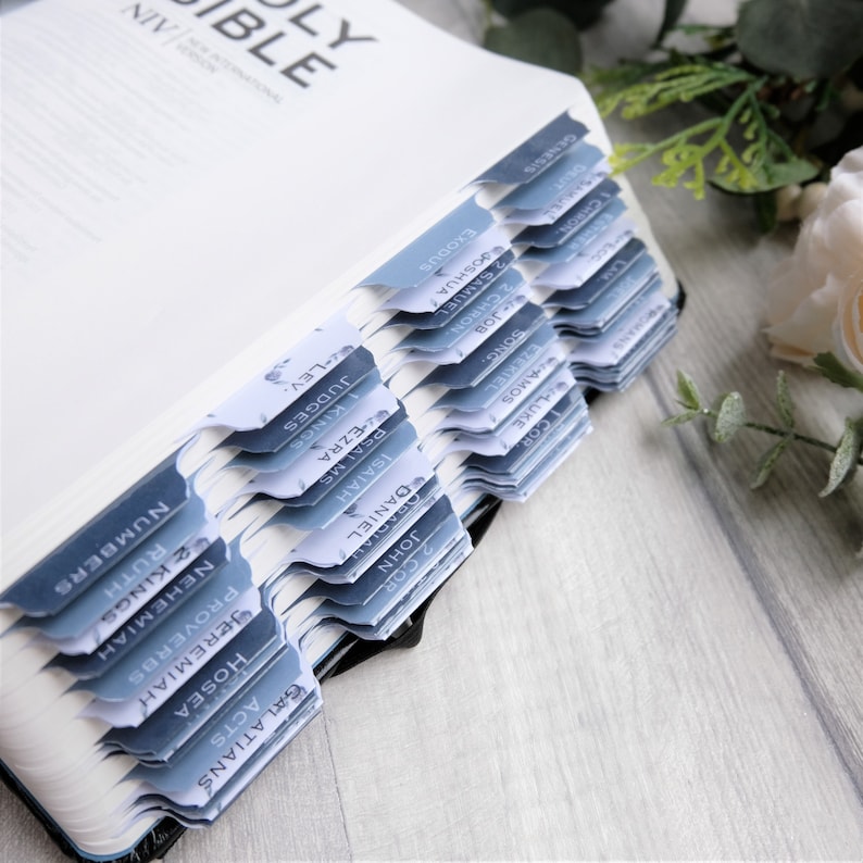 May include: A close-up of a Holy Bible with blue and white tabs marking each book of the Bible. The tabs are labeled with the names of the books, such as Genesis, Exodus, Leviticus, Numbers, Deuteronomy, Joshua, Judges, Ruth, 1 Samuel, 2 Samuel, 1 Kings, 2 Kings, 1 Chronicles, 2 Chronicles, Ezra, Nehemiah, Esther, Job, Psalms, Proverbs, Ecclesiastes, Song of Solomon, Isaiah, Jeremiah, Lamentations, Ezekiel, Daniel, Hosea, Joel, Amos, Obadiah, Jonah, Micah, Nahum, Habakkuk, Zephaniah, Haggai, Zechariah, Malachi, Matthew, Mark, Luke, John, Acts, Romans, 1 Corinthians, 2 Corinthians, Galatians, Ephesians, Philippians, Colossians, 1 Thessalonians, 2 Thessalonians, 1 Timothy, 2 Timothy, Titus, Philemon, Hebrews, James, 1 Peter, 2 Peter, 1 John, 2 John, 3 John, Jude, and Revelation.