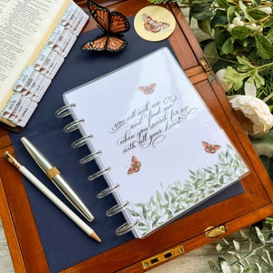 May include: A white planner with a clear cover features a quote in elegant calligraphy: "You will seek me and find me when you search for me with all your heart." The planner is decorated with green leaves and orange butterflies.