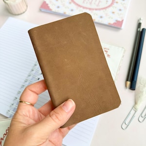 May include: A rectangular, brown leather notebook cover with a smooth, matte finish and rounded corners. The cover is plain, featuring a simple, minimalist design without any visible branding, text, or additional decorative elements on its surface.