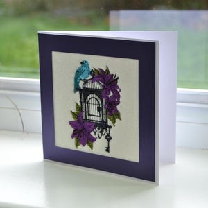 May include: Greeting card with a detailed embroidered design. The artwork features a teal bird perched atop a black birdcage, surrounded by purple flowers and green leaves. A key hangs below the cage. The card is framed in a dark purple mat.