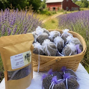 May include: A brown paper pouch labeled Blue Mount True Lavender Bud Sachets 1/2 oz next to a wooden basket filled with small white organza bags containing dried lavender buds, with additional lavender sachets arranged on a white surface.