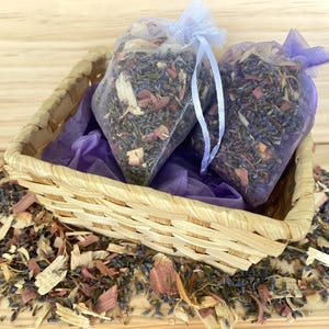 May include: Two small, clear bags filled with dried herbs and flowers, tied with white ribbons, sit in a woven basket lined with purple tissue paper. The basket is surrounded by loose herbs and wood shavings, creating a natural, rustic aesthetic.