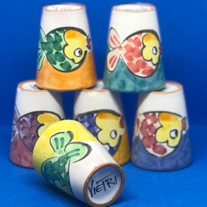May include: Six ceramic cups with a fish design. Each cup has a different color scheme and fish design. The cups are stacked on top of each other. The bottom cup has the word "Vietri" written on the bottom.