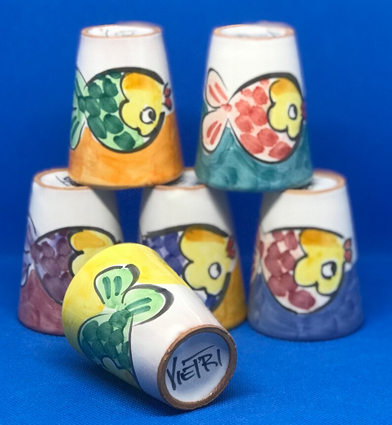 May include: Six ceramic cups with a fish design. Each cup has a different color scheme and fish design. The cups are stacked on top of each other. The bottom cup has the word "Vietri" written on the bottom.