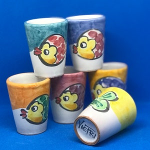 May include: Six colorful ceramic shot glasses with a fish design. Each glass has a different color and fish design. The glasses are stacked on top of each other.