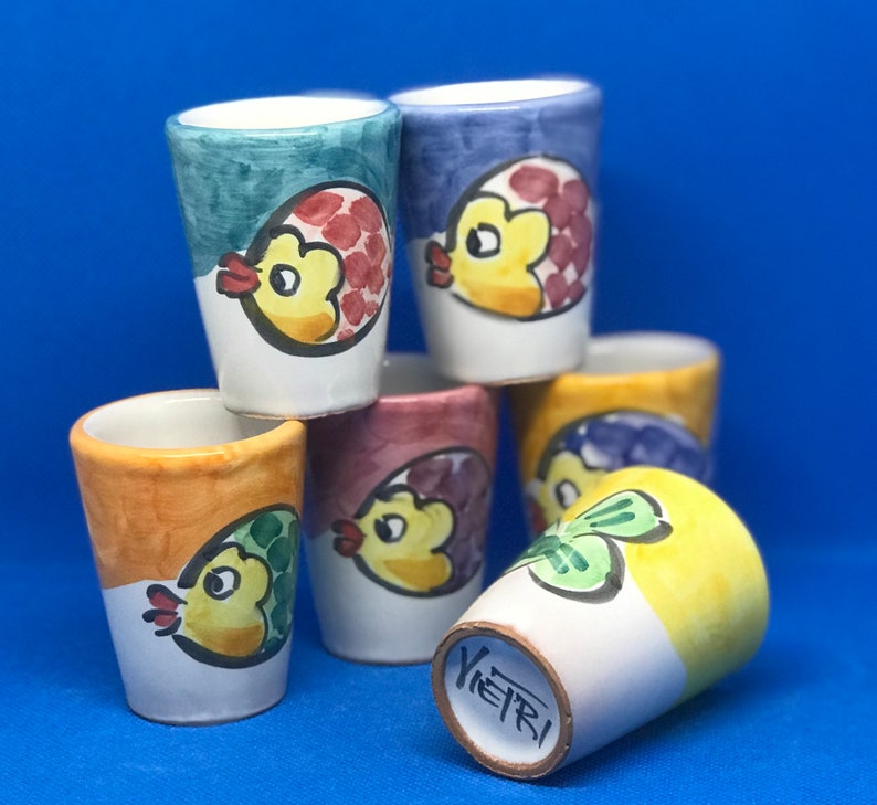 May include: Six colorful ceramic shot glasses with a fish design. Each glass has a different color and fish design. The glasses are stacked on top of each other.