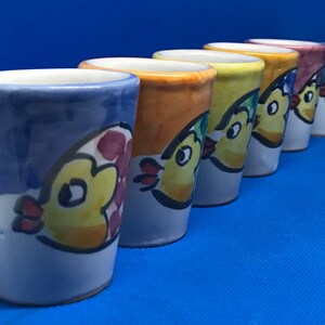 May include: Six colorful ceramic shot glasses with a whimsical design of a yellow chick with red accents. Each glass has a different color base, including blue, orange, yellow, green, and pink.