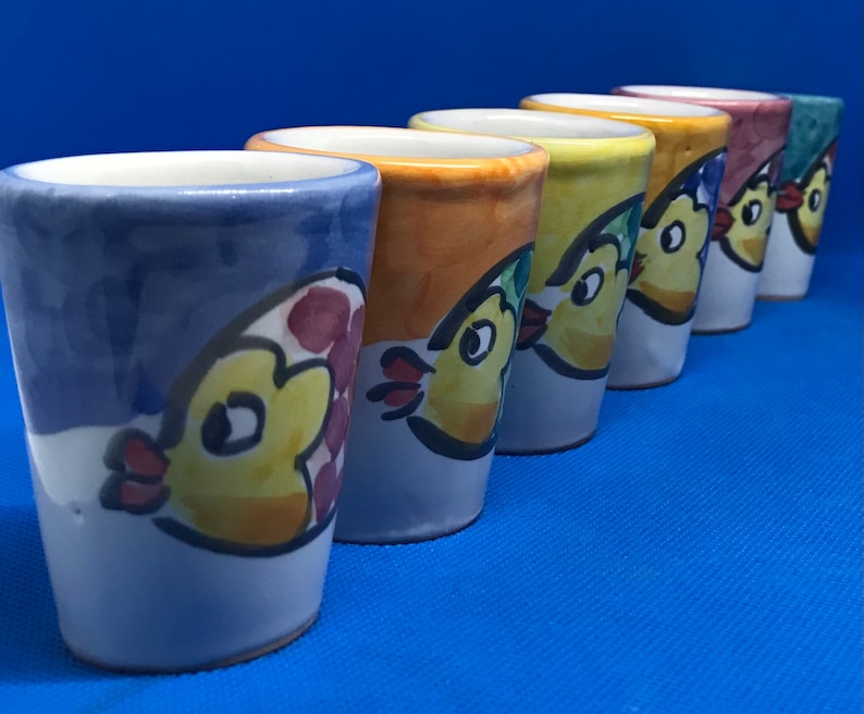 May include: Six colorful ceramic shot glasses with a whimsical design of a yellow chick with red accents. Each glass has a different color base, including blue, orange, yellow, green, and pink.