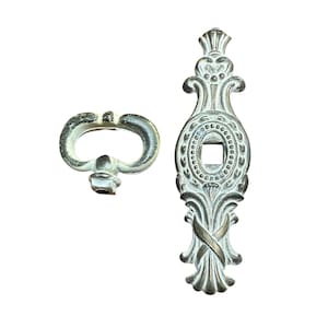 May include: Two antique-style metal cabinet hardware pieces. One is a keyhole cover with an oval center and ornate detailing. The other is a matching key with a curved handle. Both pieces have a distressed, aged finish.