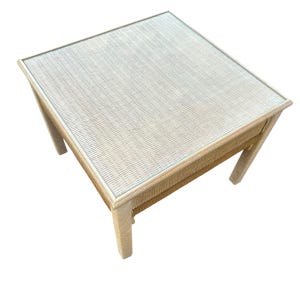 May include: Square side table with a woven beige frame and a glass top. The table has a light beige woven pattern on the top and sides. The table is approximately 24 inches wide, 24 inches long, and 20 inches high.