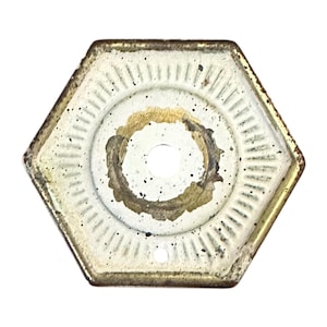 May include: Hexagonal, off-white metal object with a central hole. The object has a circular design in the center with a gold and brown ring. The edges are gold, and the surface has a distressed appearance.