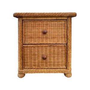 May include: A light brown wicker nightstand featuring a rectangular top with a braided edge, two stacked drawers with woven textures, and two small round wooden knobs. The piece is supported by four short, rounded woven feet at the base.