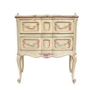 May include: A cream-colored, two-drawer chest with pink accents. The chest features ornate drawer pulls and decorative trim. The legs are curved, and the overall style is vintage. The chest is approximately 30 inches tall.
