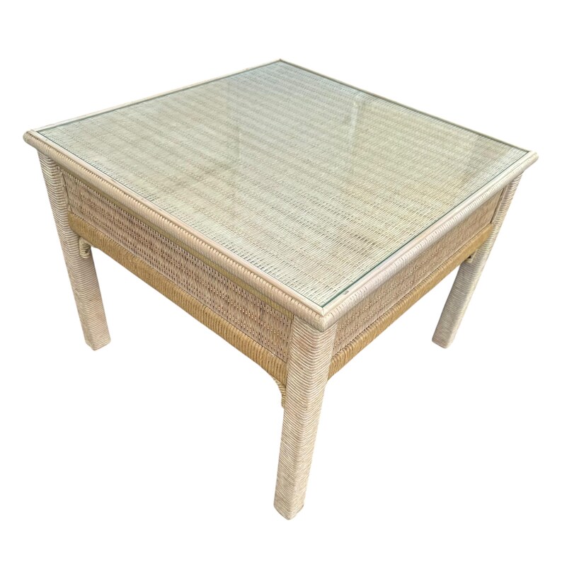 May include: Square wicker coffee table with a glass top. The table has a light beige woven design and four sturdy legs. The glass top provides a smooth surface for placing items. A stylish addition to any living room.