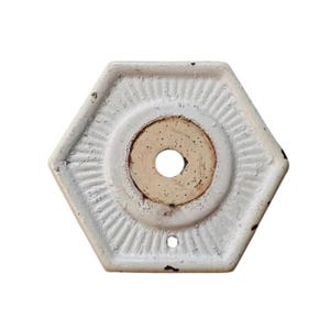 May include: A hexagonal white metal plate with a distressed finish and a central circular opening. The plate features a radiating sunburst pattern around the center and a small hole near the bottom edge, showing signs of wear and dark oxidation.