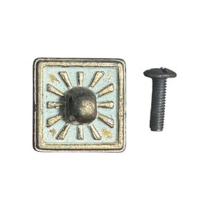May include: Square cabinet knob with a light blue center and gold sunburst design. The knob has a dark metal center and a matching screw is next to it. The knob is likely for a drawer or cabinet.