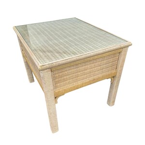 May include: A rectangular side table with a woven wicker frame and a glass top. The table is light beige in color and has four sturdy legs. The table measures approximately 24 inches long, 18 inches wide, and 20 inches high.