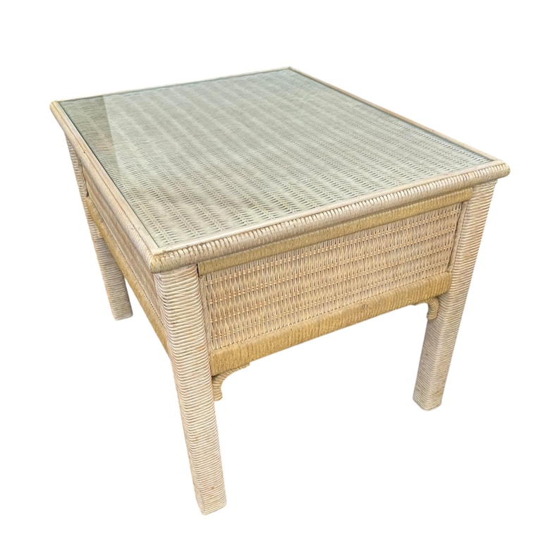 May include: A rectangular side table with a woven wicker frame and a glass top. The table is light beige in color and has four sturdy legs. The table measures approximately 24 inches long, 18 inches wide, and 20 inches high.