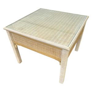May include: Square wicker coffee table with a glass top. The table has a light beige woven design and four sturdy legs. The table is approximately 24 inches wide, 24 inches long, and 18 inches high.