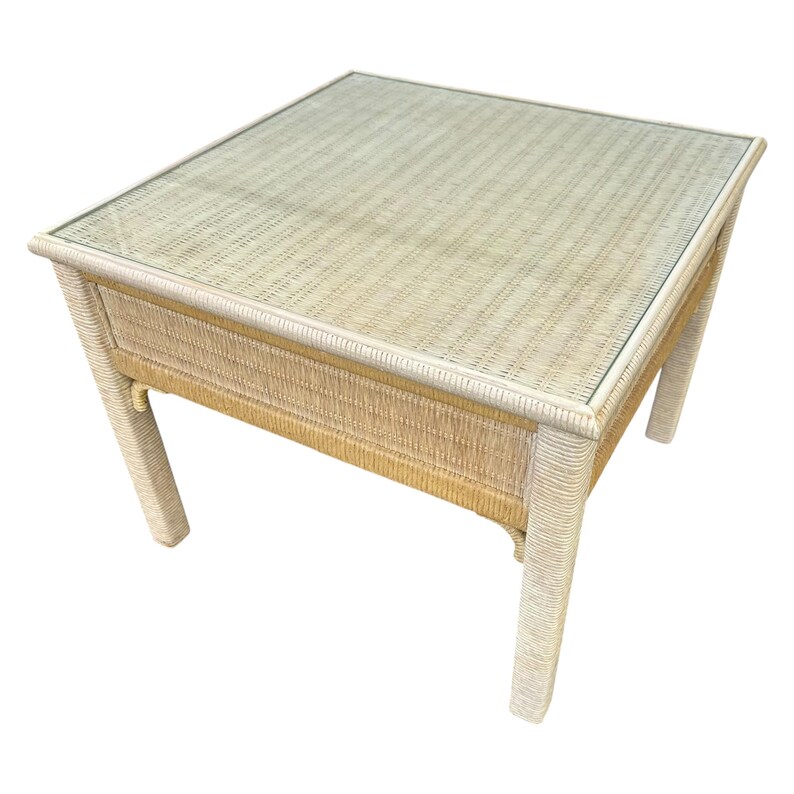 May include: Square wicker coffee table with a glass top. The table has a light beige woven design and four sturdy legs. The table is approximately 24 inches wide, 24 inches long, and 18 inches high.