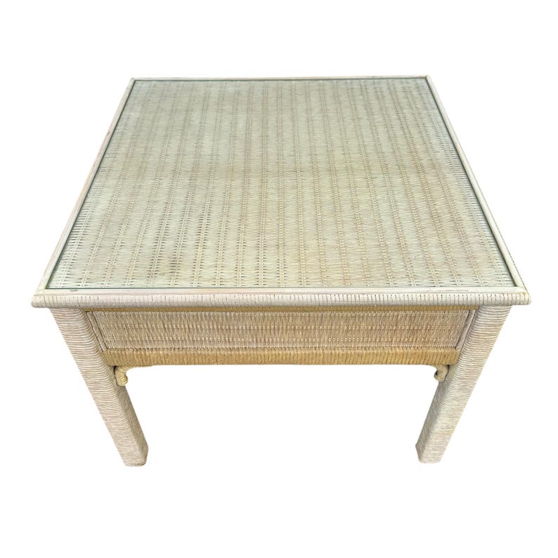 May include: Square, light beige wicker side table. The table has a woven top and sides, with a single drawer. The legs are also made of wicker. The table measures approximately 24 inches square and 20 inches high.