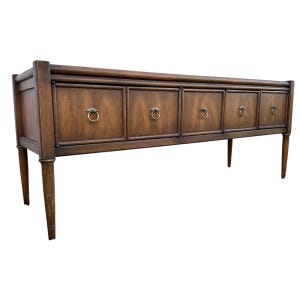 May include: A long, wooden sideboard with six drawers and ring-shaped pulls. The sideboard has a dark brown finish and sits on four slender legs.