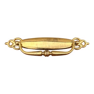 May include: A gold-toned metal drawer pull featuring an elongated, smooth central plate with decorative, openwork scroll-like ends. A curved, hinged handle with a central decorative bead hangs from the plate, providing a functional grip.