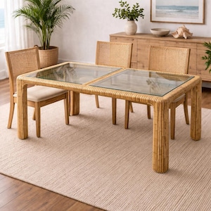 May include: A dining room set featuring a rectangular glass-top table with a natural rattan frame. Four matching rattan chairs with beige upholstered seats surround the table. The room has a neutral color palette with a woven rug and wooden furniture.