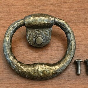 May include: A circular brass ring pull handle featuring a textured, aged finish and a central hexagonal bolt mounting plate. The hardware includes two small metal screws with rounded heads, positioned to the right of the ring pull handle.