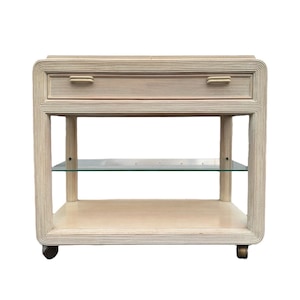 May include: A white rattan bar cart with a single drawer and a glass shelf. The cart has rounded edges and is on wheels.