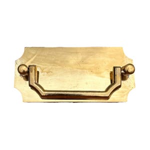 May include: A rectangular brass drawer pull featuring a polished gold-tone finish with a central drop handle. The mounting plate has notched corners and two circular attachment points where the hinged handle connects to the main metal base.