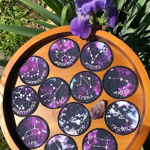 Puede incluir: Un conjunto de parches redondos con constelaciones del zodiaco sobre un fondo de galaxia púrpura y azul. Cada parche tiene un borde negro y el nombre de la constelación bordado en blanco. Los parches están colocados en una bandeja de madera con una flor morada.