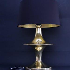 Puede incluir: Una lámpara de mesa de estilo moderno de mediados de siglo con una pantalla negra y un interior dorado. La base y el tallo son de latón pulido. La lámpara tiene una base redonda y una pantalla acampanada. Se ve un cable de alimentación negro.