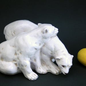 以下が含まれることがあります： 2 匹のホッキョクグマの白いセラミック彫刻。1 匹がもう 1 匹の上にあり、右側に黄色のレモンがあります。クマは光沢のある仕上げで、暗い背景に配置されています。