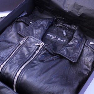 May include: A black leather jacket with a silver zipper is presented inside a black box. The jacket has a collar and the label "DOLCE & GABBANA MADE IN ITALY" is visible.