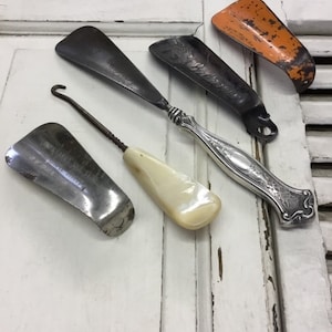 May include: Five vintage shoe horns, including a silver-plated horn with an ornate design, a horn with a mother-of-pearl handle, and two metal horns with a silver finish. The horns are arranged on a white wooden surface.