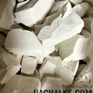 May include: A collection of irregular, matte white chalk chunks with rough, jagged edges and a powdery texture. The text UACHALKS.COM is printed in black in the lower right corner, accompanied by a small green leaf illustration.